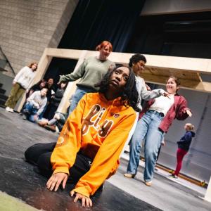 Actress sitting on the floor looking concerned wearing an orange fly sweatshirt while others are dancing behind her.