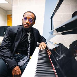 Loba Salami sits at a piano wearing a black suit and tie.