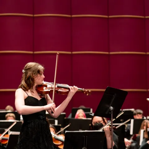 A female in the Lehigh University Philharmonic Orchestra standing up playing the violin in Baker Hall.