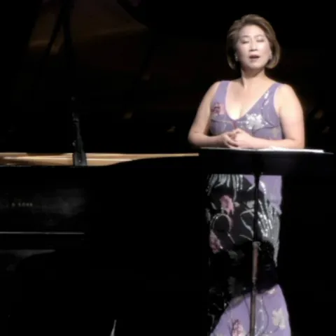 A woman wearing a purple dress singing next to a piano.