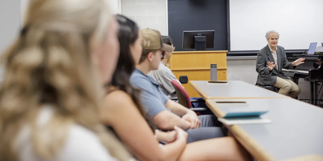Music professor sitting at piano instructing students in a classroom.