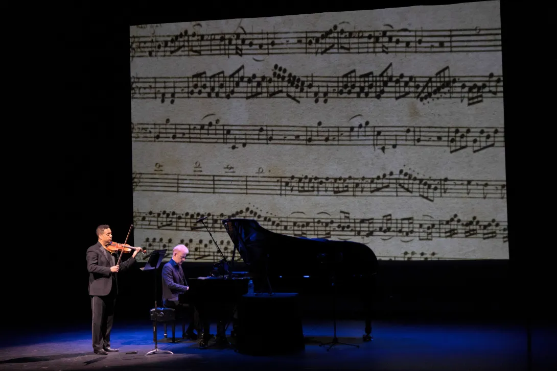 Michael Jorgensen playing the violin next to a piano player with sheet music as the background.