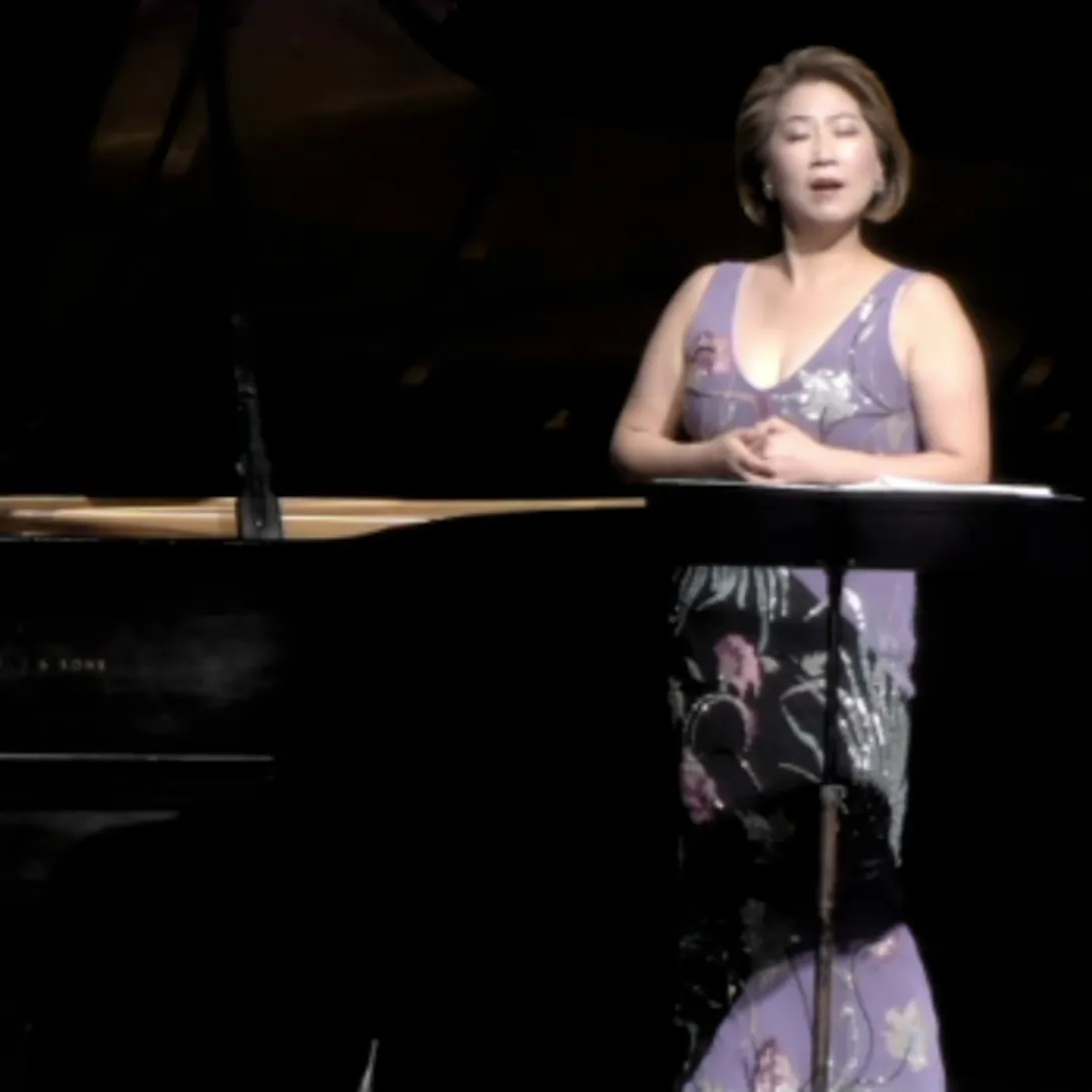 A woman wearing a purple dress singing next to a piano.