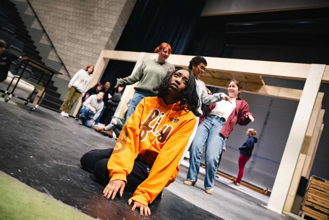 Actress sitting on the floor looking concerned wearing an orange fly sweatshirt while others are dancing behind her.